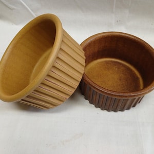 May include: Two brown ceramic ramekins with a ribbed exterior. One ramekin is tilted, revealing the interior. The other ramekin is upright. The ramekins are likely for baking or serving individual portions of food.