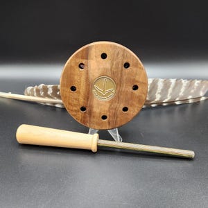 Puede incluir: Un llamador de juego de madera con un cuerpo circular marrón oscuro con ocho agujeros y un emblema de latón. Un percutor de madera de color claro con un eje verde y marrón descansa a su lado. Una pluma está en el fondo.