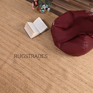May include: A natural jute rug with a subtle striped pattern. The rug is beige in color and has a slightly rough texture. The rug is shown with a book and a beanbag chair.