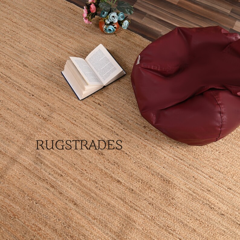 May include: A natural jute rug with a subtle striped pattern. The rug is beige in color and has a slightly rough texture. The rug is shown with a book and a beanbag chair.