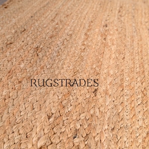 Puede incluir: Una alfombra tejida de color tostado con una textura trenzada gruesa hecha de fibras naturales. La superficie presenta un patr&oacute;n repetitivo de bucles entrelazados. El texto RUGSTRADES est&aacute; impreso en letras may&uacute;sculas negras sin remates en el centro.