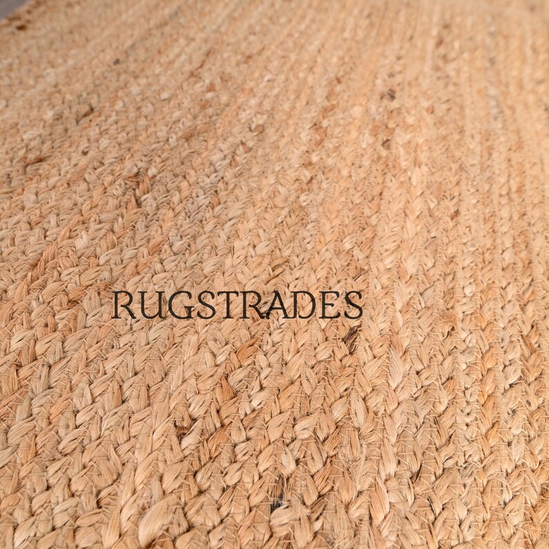 Puede incluir: Una alfombra tejida de color tostado con una textura trenzada gruesa hecha de fibras naturales. La superficie presenta un patr&oacute;n repetitivo de bucles entrelazados. El texto RUGSTRADES est&aacute; impreso en letras may&uacute;sculas negras sin remates en el centro.