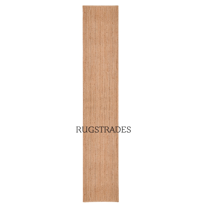 Puede incluir: Una alfombra de pasillo larga y rectangular hecha de fibras de yute natural de color tostado con una superficie tejida y texturizada. La alfombra presenta un tono marr&oacute;n claro uniforme e incluye el texto negro RUGSTRADES en el centro.