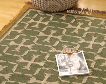 May include: Green and beige patterned rug with a geometric design. A cocktail glass and a magazine rest on the rug. A woven ottoman and a wooden floor are in the background. The rug has a brown border and fringe.