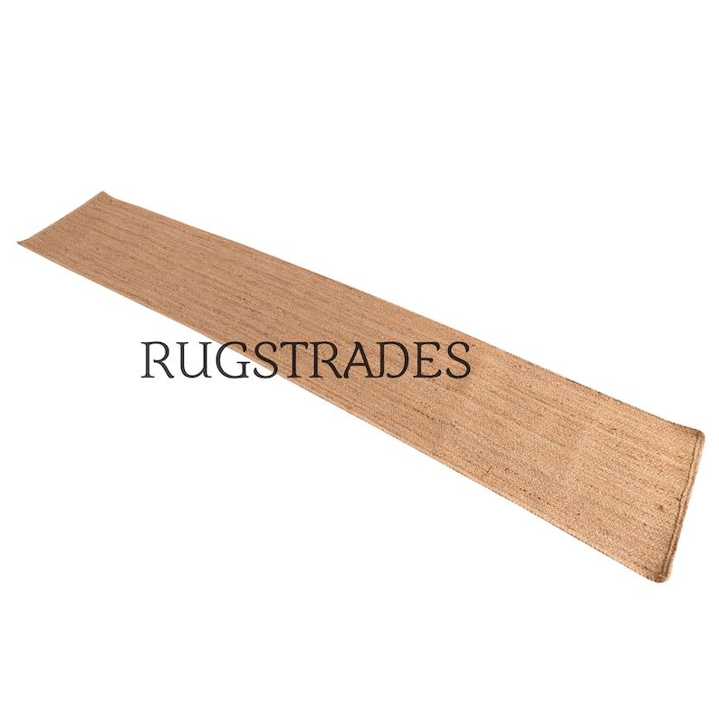 Puede incluir: Una alfombra de pasillo larga y rectangular hecha de fibras tejidas de color tostado natural con una superficie texturizada visible. El texto RUGSTRADES est&aacute; impreso en letras may&uacute;sculas negras en el centro, sobre un fondo blanco.