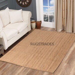 May include: A natural fiber rug in a light brown color with a woven texture. The rug is laid out on a wooden floor in a living room setting. The text "RUGSTRADES" is visible in the center of the rug.