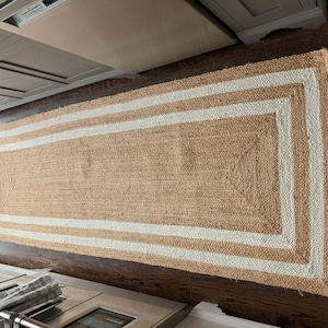 以下が含まれることがあります： 白い縁取りのある天然繊維の敷物。敷物はジュート製で、織り込まれた質感があります。