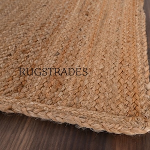 Puede incluir: Una alfombra de yute trenzado de color tostado con una textura tejida gruesa y un borde visible. La palabra RUGSTRADES est&aacute; impresa en letras may&uacute;sculas negras sin remates en el centro de la superficie de la alfombra, mostrando el material de fibra.
