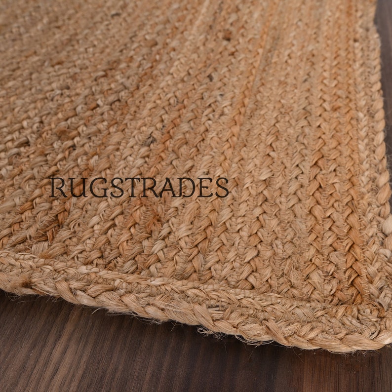 Puede incluir: Una alfombra de yute trenzado de color tostado con una textura tejida gruesa y un borde visible. La palabra RUGSTRADES est&aacute; impresa en letras may&uacute;sculas negras sin remates en el centro de la superficie de la alfombra, mostrando el material de fibra.