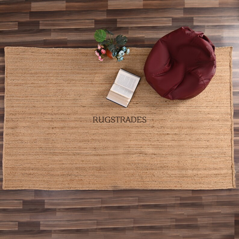 May include: A natural jute rug with a subtle striped pattern. The rug is beige and has a slightly rough texture. A red beanbag chair and a book are placed on the rug.