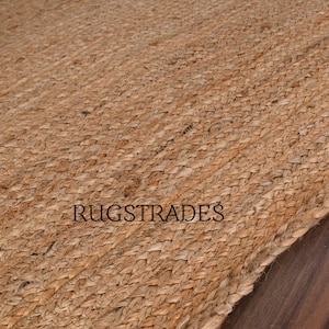 Puede incluir: Una alfombra tejida de color tostado con un patr&oacute;n trenzado y texturizado hecho de fibras naturales. La superficie presenta una estructura de tejido diagonal repetitivo. El texto RUGSTRADES est&aacute; impreso en letras may&uacute;sculas negras sin remates en el centro.