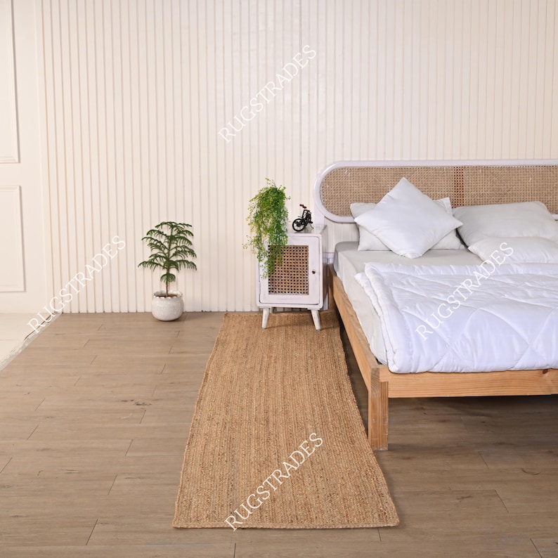 Puede incluir: Una alfombra pasillera rectangular de fibras naturales con textura tejida sobre un suelo de madera junto a una cama con edred&oacute;n y almohada blancos, y una peque&ntilde;a mesita de noche con frente de rejilla y una planta verde encima.