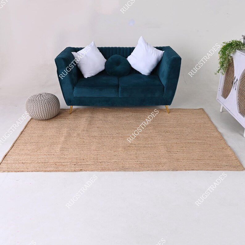 Pu&ograve; includere: Un tappeto in juta marrone chiaro con un divano in velluto turchese con due cuscini bianchi e un pouf rotondo grigio di fronte.