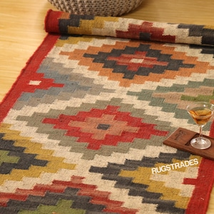 以下が含まれることがあります： 赤、オレンジ、グレー、緑、黄色の幾何学模様の長方形の丸められたラグ。ラグには赤い縁があり、右下隅に「RUGSTRADES」という文字が見えます。