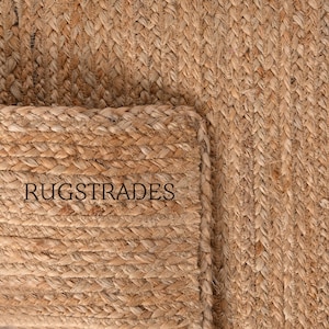 Puede incluir: Una alfombra de yute tejida de color tostado con una textura trenzada y una esquina doblada. La palabra RUGSTRADES est&aacute; impresa en letras may&uacute;sculas negras sobre la superficie de la alfombra, mostrando la fibra natural y el patr&oacute;n de tejido.