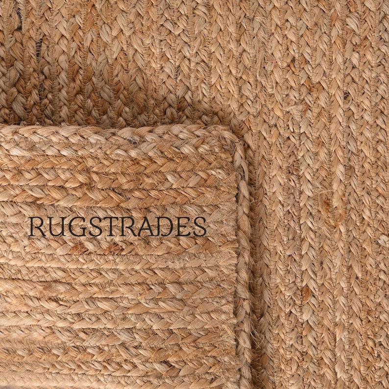 Puede incluir: Una alfombra de yute tejida de color tostado con una textura trenzada y una esquina doblada. La palabra RUGSTRADES est&aacute; impresa en letras may&uacute;sculas negras sobre la superficie de la alfombra, mostrando la fibra natural y el patr&oacute;n de tejido.