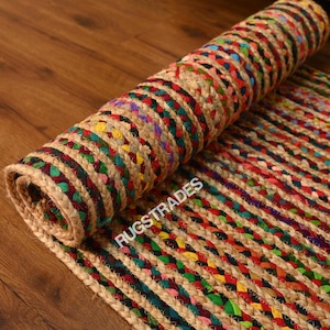 May include: Rolled jute rug with a braided design. The rug features alternating stripes of natural jute and colourful braided sections in red, green, yellow, and purple. The text "RUGSTRADES" is visible on the rug.