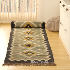 May include: A rolled-up, rectangular rug with a geometric pattern in shades of green, yellow, and black. The rug has a dark gray border and fringe at the end. The text "RUGSTRADES" is visible on the rug.