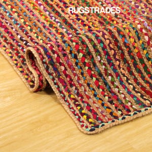 May include: A multi-coloured braided jute rug with red, blue, green, and yellow highlights. The rug is partially rolled up, showing its textured surface and the "RUGSTRADES" text. The rug is placed on a light brown wooden floor.