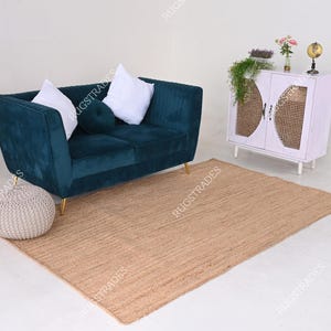 Pu&ograve; includere: Un divano in velluto verde acqua con due cuscini bianchi e un cuscino marrone &egrave; posizionato su un tappeto in juta naturale. Uno sfondo con un mobile bianco con una porta in rattan intrecciato.