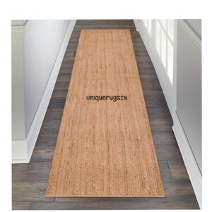 Puede incluir: Una alfombra de pasillo de fibra natural de color marrón claro. La alfombra está hecha de yute y tiene una textura tejida. La alfombra está colocada en un pasillo con paredes blancas y suelo de madera. El texto "uniquerugsIN" es visible en la alfombra.