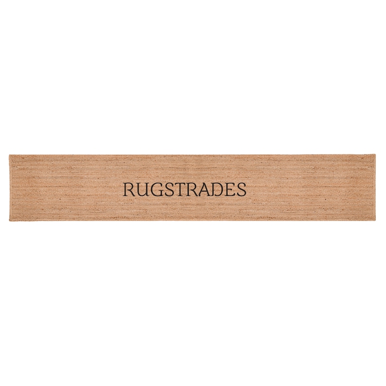 Puede incluir: Una alfombra rectangular tipo pasillo hecha de fibras tejidas de color canela natural con una superficie texturizada. La palabra RUGSTRADES est&aacute; impresa en letras may&uacute;sculas negras en el centro de la alfombra, con un tejido denso y marr&oacute;n.
