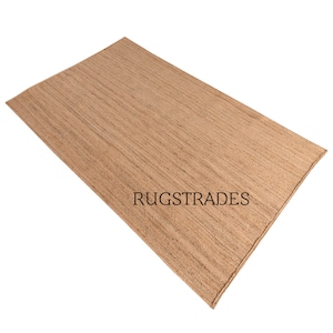 May include: A natural fiber rug made from jute, a sustainable and durable material. The rug has a light brown color and a woven texture. The rug is shown in a flat lay, with the text "RUGSTRADES" printed on the surface.