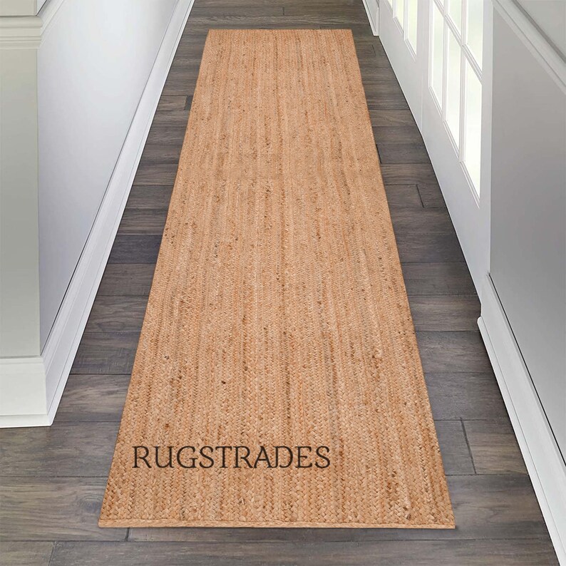 May include: A natural fiber runner rug in a light brown color. The rug is made of jute and has a woven texture. The rug is placed on a hardwood floor in a hallway. The text "RUGSTRADES" is printed on the rug.