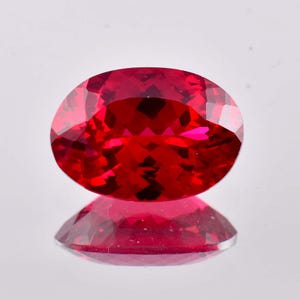 May include: A faceted, oval-shaped, ruby gemstone. The gem is a deep, vibrant red color with internal reflections. The stone is set against a reflective surface, creating a mirrored effect.