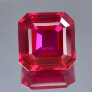 May include: A vibrant red gemstone with a square emerald cut. The gemstone is faceted and has a high polish.