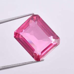 May include: A faceted, rectangular pink gemstone held by silver tweezers. The gem has a vibrant, transparent color and a symmetrical cut, reflecting light. The gemstone is a vivid shade of pink, with sharp edges and a polished surface.