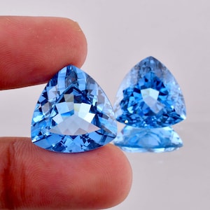 May include: Two triangular, faceted, light blue gemstones. One is held between fingers, the other is in the background with its reflection visible. The gems have a brilliant, sparkling appearance, suggesting they are cut with precision.