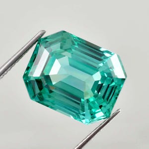 Op de afbeelding: Een gefacetteerde, smaragdgeslepen edelsteen in een levendige turquoise kleur. De edelsteen wordt vastgehouden door een zilveren pincet, waardoor de geometrische facetten en interne reflecties worden benadrukt. De rechthoekige vorm wordt geaccentueerd door de trapsgewijze snede.