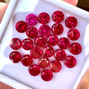 May include: A selection of round, faceted rubies in shades of red and pink. The gemstones are presented in a white tray, highlighting their sparkle and clarity. The rubies are of a consistent size and shape.