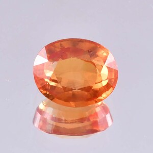 May include: An oval-cut, orange gemstone with a faceted surface. The gem is a vibrant shade of orange, with a reflective surface below. The gemstone is likely a sapphire or topaz, suitable for jewelry making.