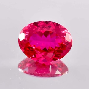 May include: A vibrant pink oval-shaped gemstone with a faceted cut. The gemstone is reflecting light, showcasing its brilliance.