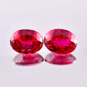May include: Two oval-cut, faceted rubies with a vibrant red hue. The gemstones are displayed on a reflective surface, showcasing their brilliance and clarity. The rubies are a deep, rich red, with visible internal structures.