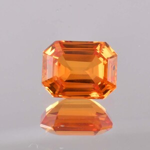 May include: A faceted, rectangular-cut gemstone with an orange hue. The gem is highly reflective, with multiple facets creating a brilliant display of light. The gemstone is set against a reflective surface, creating a mirrored effect.