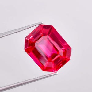 May include: A vibrant, faceted ruby gemstone held by silver tweezers. The gem has a rectangular emerald cut with a deep red hue and internal reflections, set against a white background. The tweezers are visible at the top and bottom of the gem.