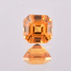 May include: A faceted, square-cut, orange gemstone with a reflective surface. The gem is a vibrant shade of orange, with multiple facets that catch the light. Below the gem is a triangular reflection of the gem.