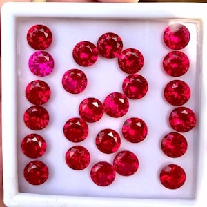 Könnte beinhalten: Eine Sammlung von 20 runden, roten Edelsteinen, die in einer weißen quadratischen Schachtel präsentiert werden. Die Edelsteine sind in einem Muster angeordnet, einige in Reihen und andere in einer lockeren Gruppe.