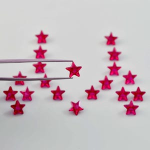 Op de afbeelding: Een set van 15 roze stervormige strasssteentjes, waarvan er één met een pincet wordt vastgehouden. De strasssteentjes zijn in een patroon op een wit oppervlak gerangschikt.