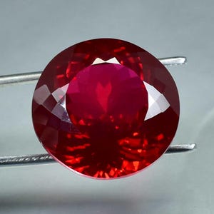 May include: A faceted, round-cut ruby gemstone. The gem is a deep, vibrant red color, held by silver tweezers against a light gray background. The facets create a brilliant display of light and shadow.