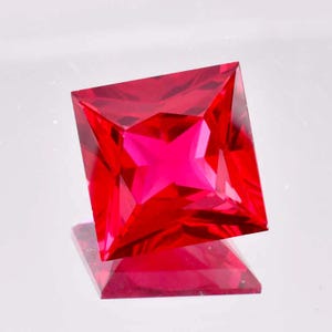 May include: A vibrant red square-shaped gemstone with a faceted cut. The gemstone is reflecting light, creating a sparkling effect.