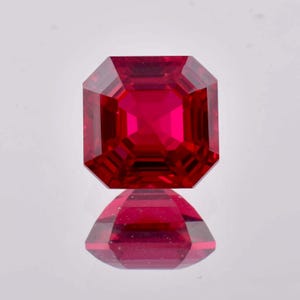 May include: A deep red gemstone with a square emerald cut. The gemstone is faceted and has a high polish.