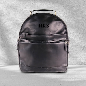 May include: Black leather backpack with a top handle and multiple pockets. The backpack has the initials "HKS" embroidered on the front.