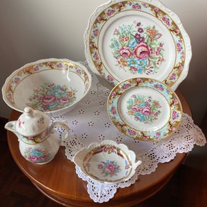 Könnte beinhalten: Ein Vintage-Porzellan-Dinner-Set mit floralen Mustern in Rosa-, Blau- und Gelbtönen. Das Set beinhaltet einen großen Speiseteller, einen kleineren Salatteller, eine Suppenschüssel, einen Milchkännchen und eine kleine Schale. Die Teile sind mit goldenen Verzierungen und aufwendigen Blumenmustern verziert.