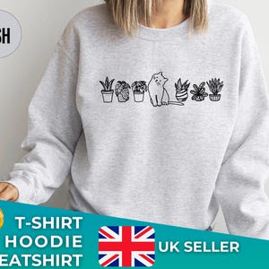 May include: A grey sweatshirt featuring a black and white illustration of a cat sitting in front of a row of potted plants. The text "ASH" is in a white circle in the top left corner of the image. The text "T-SHIRT HOODIE SWEATSHIRT UK SELLER" is at the bottom of the image.