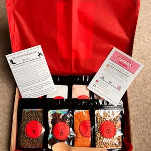 May include: A cardboard box containing a sensory play kit with red tissue paper, a wooden spoon, and several black pouches filled with various dried ingredients. Two informational cards are included, one titled Recommended Potion Recipe and the other Potions by Post.