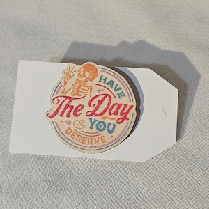 Könnte beinhalten: Ein rundes Holzschild mit einem Skelett, das ein Peace-Zeichen zeigt. Das Schild lautet "Have the Day You Deserve."